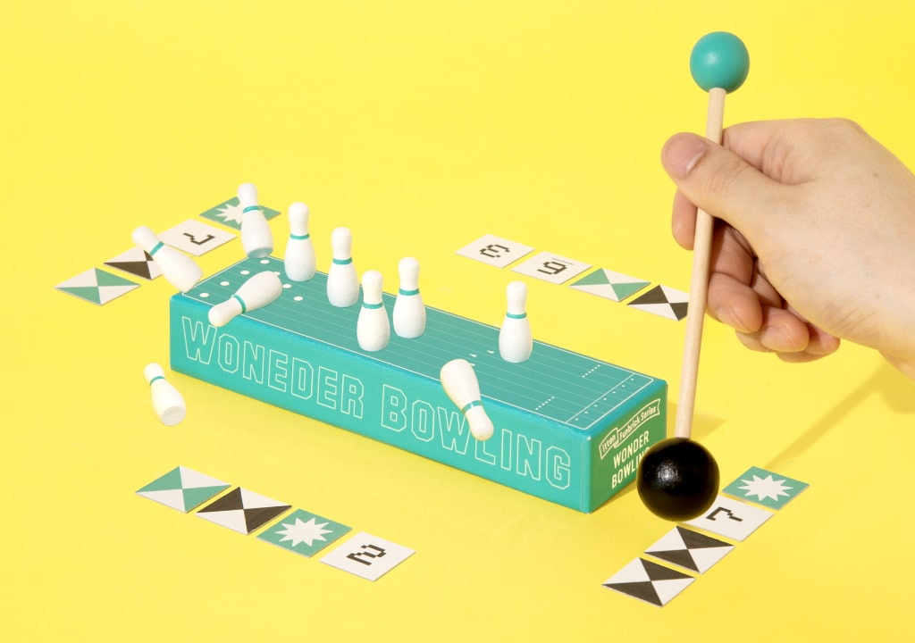 Game Shelf Wonder Bowling žaidimas – medinė lazda smūgiuoja boulingo kamuoliu į kėglius, geltonas fonas.